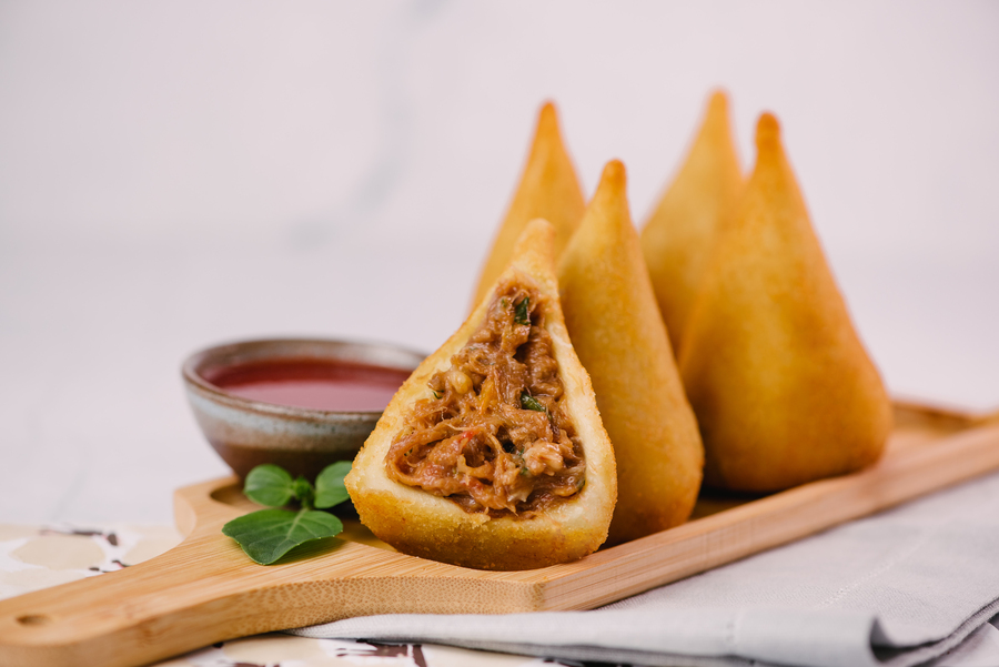 Coxinhas triangulares recheadas, destacando a origem da coxinha, servidas com molho vermelho em uma tábua de madeira.